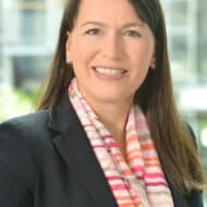 Porträt von Annette Elzenheimer in Blazer mit gestreiftem Schal, lächelnd vor unscharfem, modernen Hintergrund.