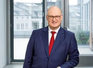Gimmy-Marc Andre-sit-ppt Mann in Anzug mit Brille steht vor einem Fenster, lächelt, moderne Büroumgebung im Hintergrund.