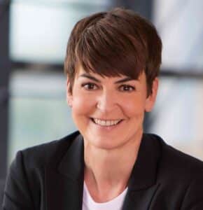 Silvia_Hänig_web bearb c iKOM Porträt einer lächelnden Frau mit kurzem, braunem Haar, in einem schwarzen Blazer vor einem unscharfen Hintergrund.