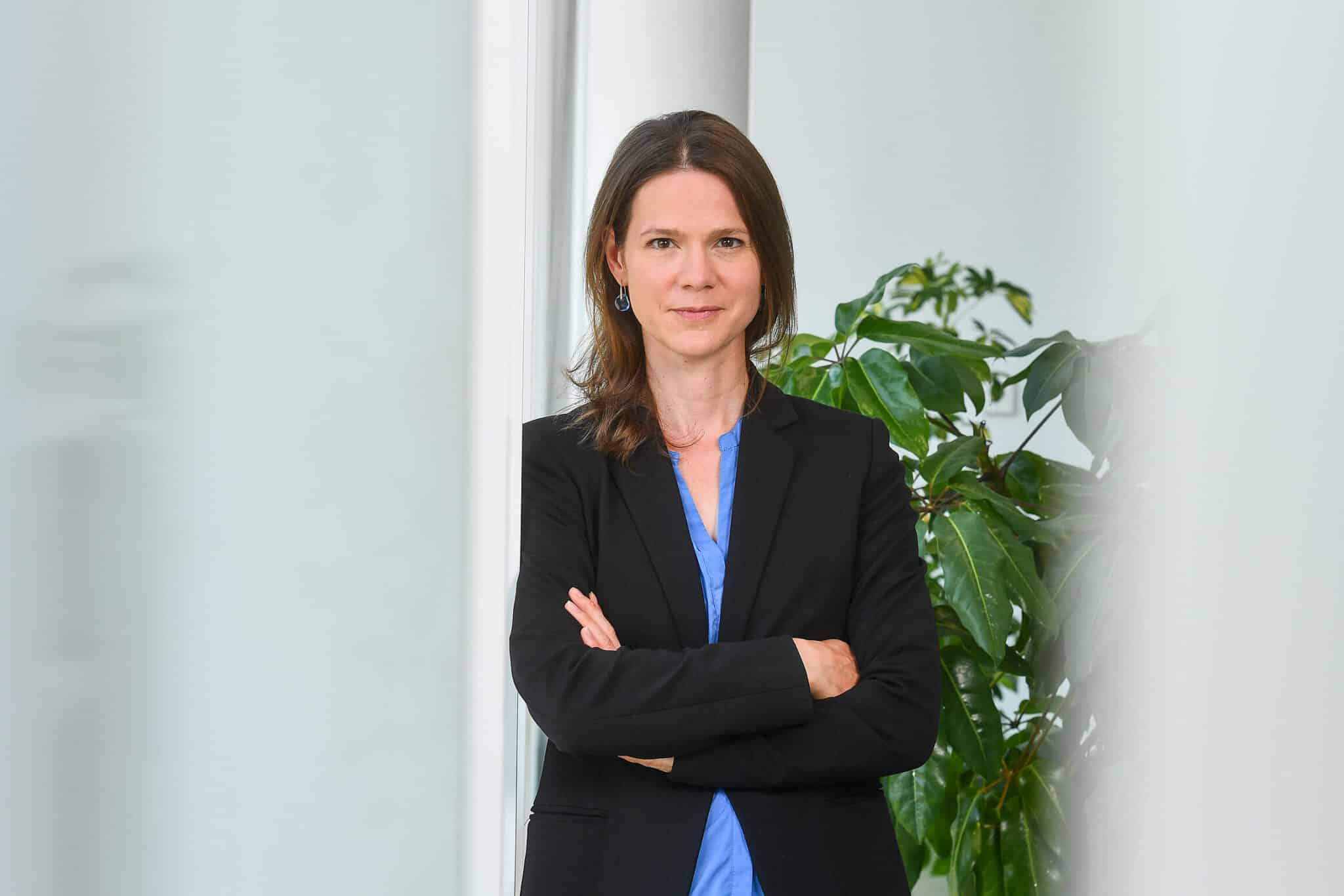 Dr. Julia Klinger steht mit verschränkten Armen vor einer Pflanze, trägt einen schwarzen Blazer und hat eine freundliche...