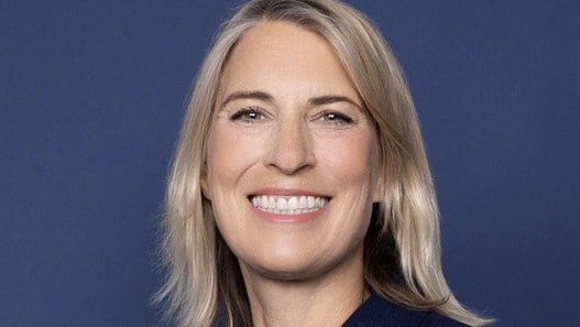 Porträt einer lächelnden Frau mit blonden Haaren vor einem blauen Hintergrund.
