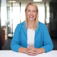 Frau mit blonden Haaren und blauer Jacke sitzt an einem Tisch und lächelt, Hintergrund mit modernen Büromöbeln.
