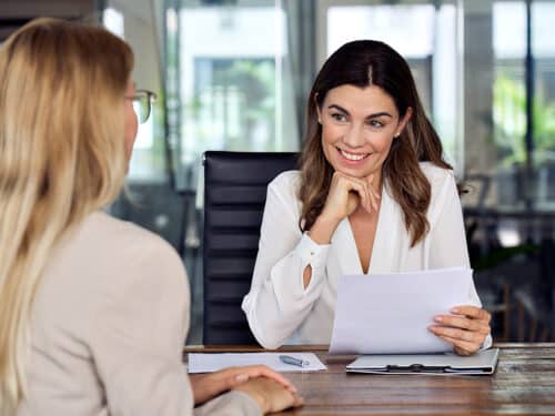 Zwei Frauen in einem modernen Büro, eine lächelt und hält Dokumente, die andere hört aufmerksam zu.
