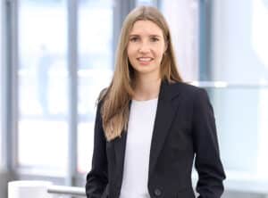 Burger-Larissa-sit-ppt Frau mit langen, blonden Haaren in schwarzem Blazer steht lächelnd vor großen Fenstern.