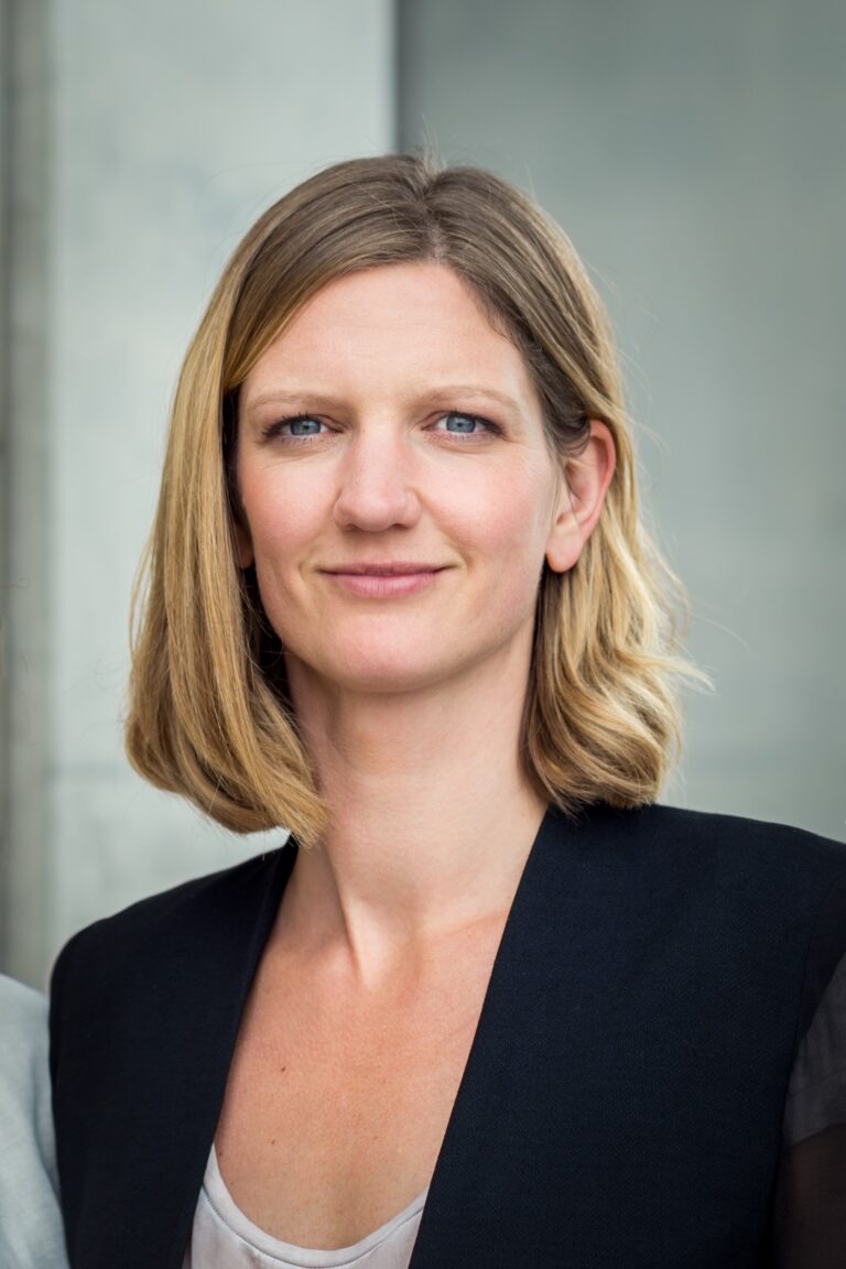 Portrait of a woman with shoulder-length blonde hair wearing a black blazer, standing beside a light wall outdoors.