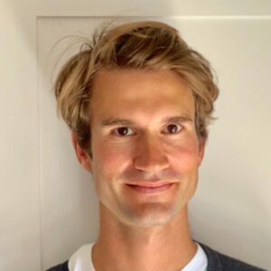 Porträt eines lächelnden Mannes mit blonden, lockigen Haaren vor einem neutralen Hintergrund.