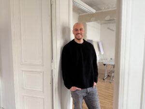 Florian Feltes c Zortify Mann mit Glatze und schwarzem Pullover steht lächelnd in einem hellen Raum mit Holzfußboden und Möbeln im Hintergrund.