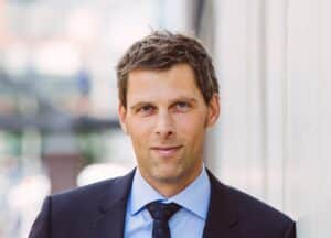 Florian Tietjen c Kick Consulting Mann in Anzug mit Krawatte, lächelt vor einer hellen Wand, unscharfer Hintergrund mit urbanem Ambiente.