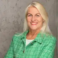 Porträt einer lächelnden Frau mit langen, blonden Haaren, die einen grünen Blazer trägt, vor einer grauen Wand.
