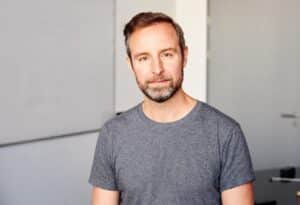 markus albers c Patrick Desbrosses Mann mit Bart in grauem T-Shirt, lächelt leicht vor neutralem Hintergrund.