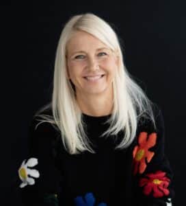 ConnyThalheim c Ishtar Najjar Lächelnde Frau mit langen, blonden Haaren in einem bunten, blumigen Pullover vor einem dunklen Hintergrund.