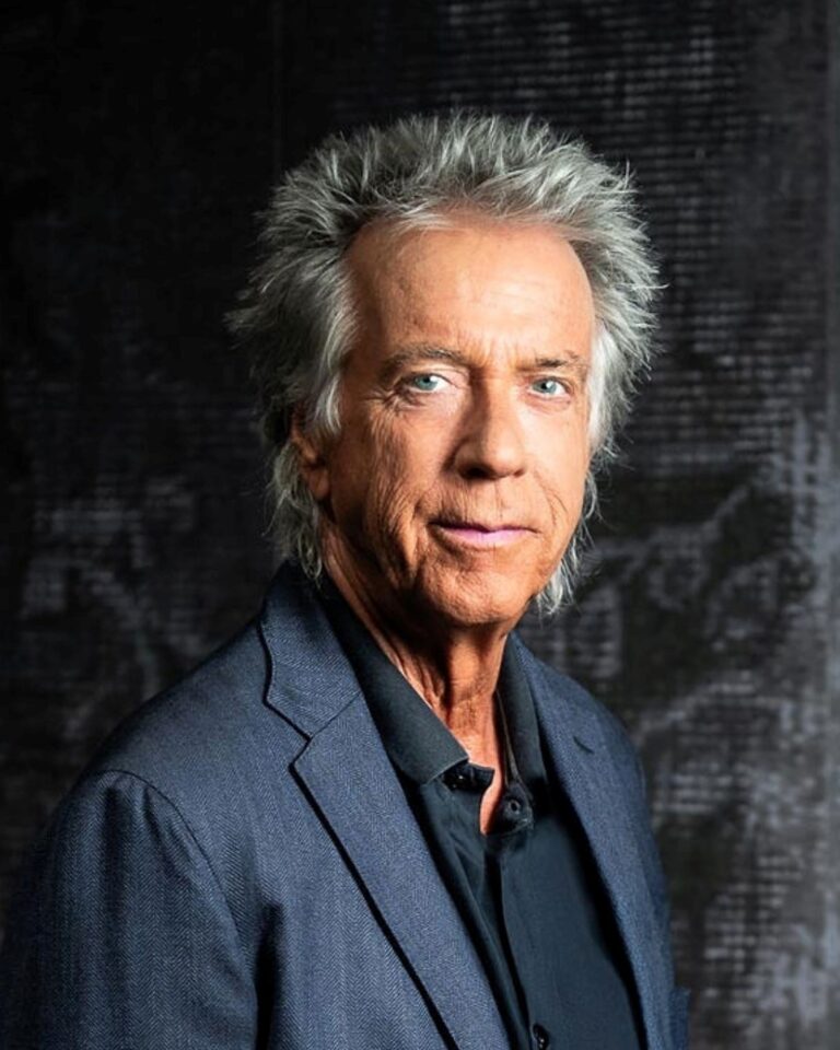 Porträt eines älteren Mannes mit grauen Haaren, in einem blauen Anzug und schwarzem Hemd, vor einem dunklen Hintergrund.