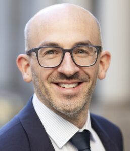 Hartmann Jan_CK_8965_Portrait_beschnitten Porträt eines lächelnden Mannes mit Glatze, Brille und Anzug, vor unscharfem Hintergrund.