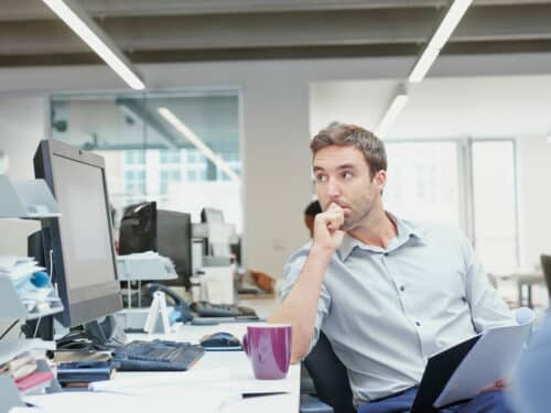 Man at an office desk thinking, resting his chin on his hand, with a computer monitor and paperwork nearby.