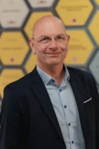 HelmutFeldmeier1 Mann mit Glatze und Brille, trägt einen dunklen Anzug und ein hellblaues Hemd, vor einem geometrischen Hintergrund.