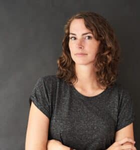 Porträt einer Frau mit lockigem Haar, die in einem grauen T-Shirt vor einem dunklen Hintergrund steht.