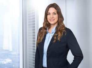KrupaSoltane-Janine-sit-ppt Frau mit langen, lockigen Haaren in einem Anzug steht vor einem Fenster mit natürlichem Licht.