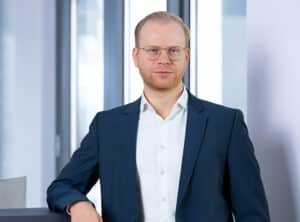 Adam-Niklas-sit-ppt Mann mit Brille in Anzug steht an einem Tisch, lächelt und schaut in die Kamera, mit Fenstern im Hintergrund.
