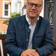 Mann mit Brille und Anzug sitzt an einem Tisch, lächelt, im Hintergrund ein Gebäude mit Fenstern und Pflanzen.