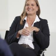 Frau mit langen Haaren in einem Anzug sitzt lächelnd und spricht, Hände gefaltet, in einem modernen Büro.