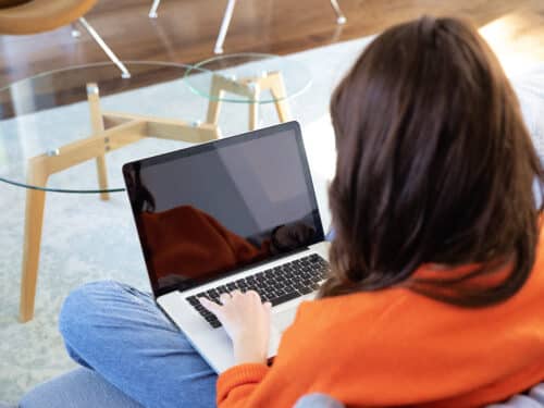 Person in an orange hoodie types on a laptop resting on their lap near a glass coffee table.