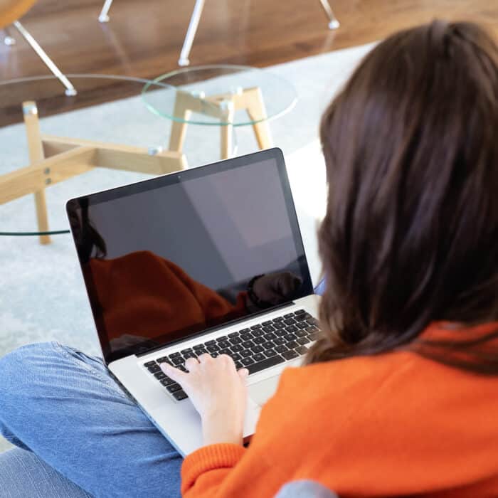 Person in an orange hoodie types on a laptop resting on their lap near a glass coffee table.