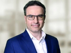 Insam_Alexander_I_ohne-Krawatte_300dpi_cmyk Porträt eines Mannes mit Brille, in einem blauen Anzug und weißem Hemd, vor unscharfem Hintergrund.