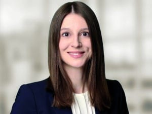 Lahmer_Sarah_300dpi_cmyk Porträt einer lächelnden Frau mit langen, glatten Haaren, in einem blauen Blazer, vor einem unscharfen Hintergrund.