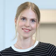 Porträt einer lächelnden Frau mit blonden Haaren, die ein gestreiftes Oberteil trägt, vor einem hellen Hintergrund.