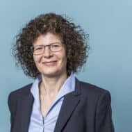 Frau mit lockigem Haar und Brille, trägt einen Blazer, lächelt vor einem blauen Hintergrund.