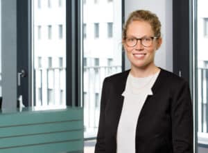 Reuter-Wiebke-sit-ppt Frau mit Brille und blonder Haarfrisur in einem modernen Büro mit großen Fenstern. Sie trägt einen schwarzen Blazer.