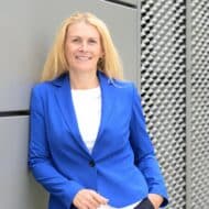 Frau mit langen blonden Haaren in blauer Blazerjacke steht lächelnd an einer grauen Wand mit strukturiertem Hintergrund.