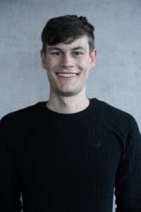 Junger Mann mit kurzem Haar, lächelt in einem schwarzen Pullover vor einer grauen Wand.