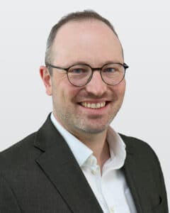 Porträt eines lächelnden Mannes mit Brille, in einem dunklen Anzug und weißem Hemd, vor einem hellen Hintergrund.