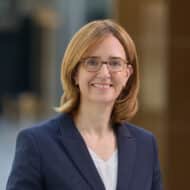 Porträt einer Frau mit blonden Haaren, Brille und dunklem Blazer, lächelnd vor unscharfem Hintergrund.