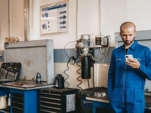 Mann in blauer Arbeitskleidung steht in einer Werkstatt und schaut auf sein Smartphone. Werkzeug und Maschinen im Hintergr...