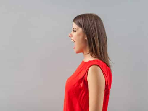 Brunette Frau in rotem Shirt, seitlich, schreit aggressiv, vor grauem Hintergrund.