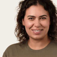 Porträt von Jennifer Weihs mit lockigem, braunem Haar, lächelnd, in einem grauen T-Shirt vor neutralem Hintergrund.